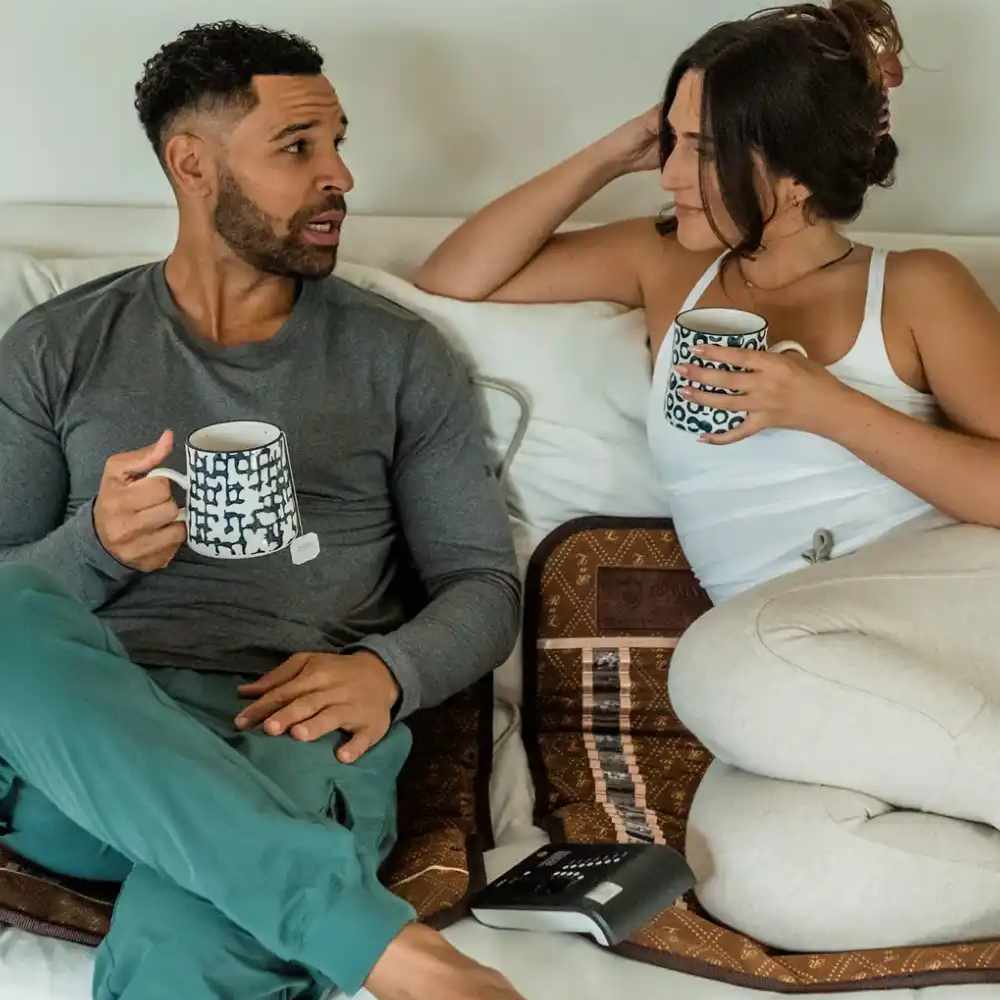 Two people sitting on BioMat Mini portable infrared therapy mats on a sofa with mugs and digital controller visible