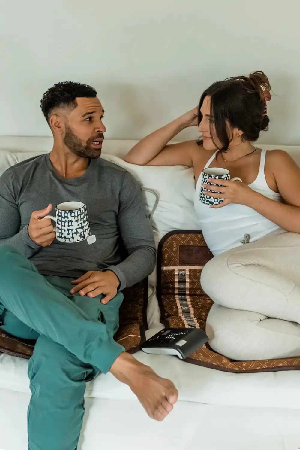 Two people sitting on BioMat Mini portable infrared therapy mats on a sofa with mugs and digital controller visible
