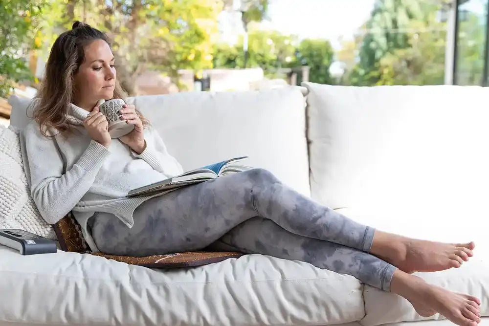 Person using the BioMat Mini portable infrared therapy mat on a couch while holding a mug and reading a book