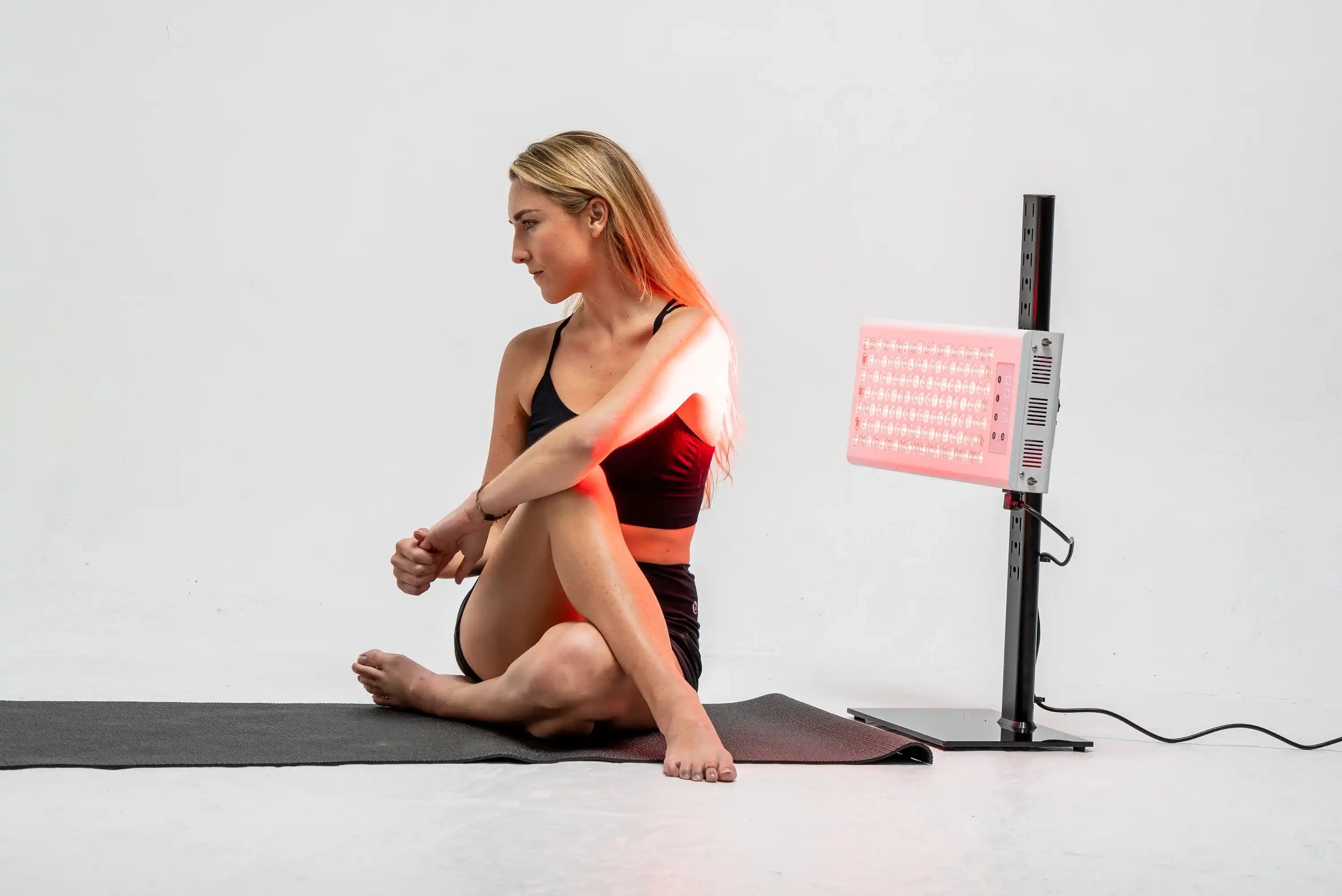 Person seated on a yoga mat beside the RedRush 400 PULSE targeted red and near-infrared light panel