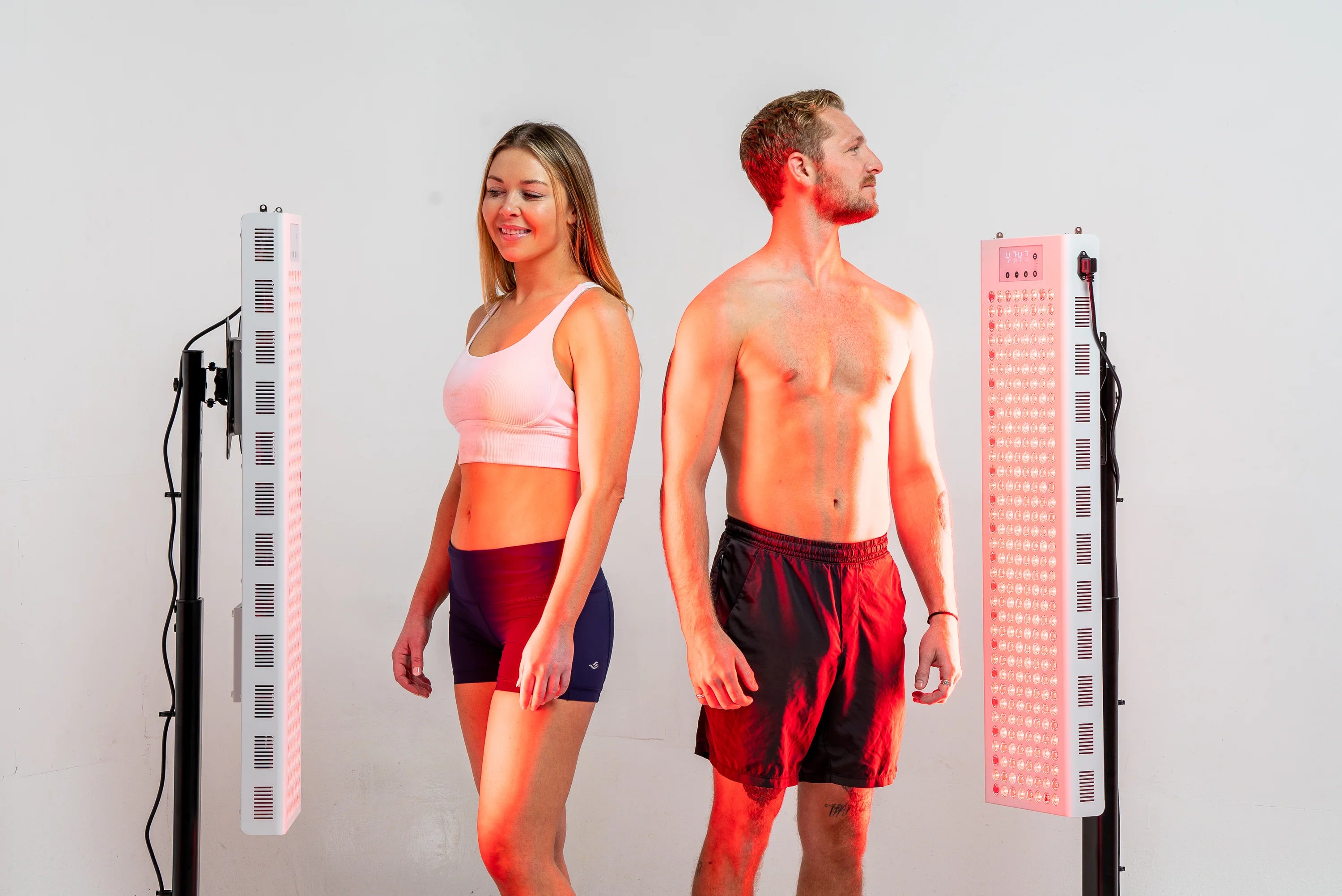 Two people standing between the illuminated panels of the RedRush 840 PULSE Dual two-panel red and near-infrared light system