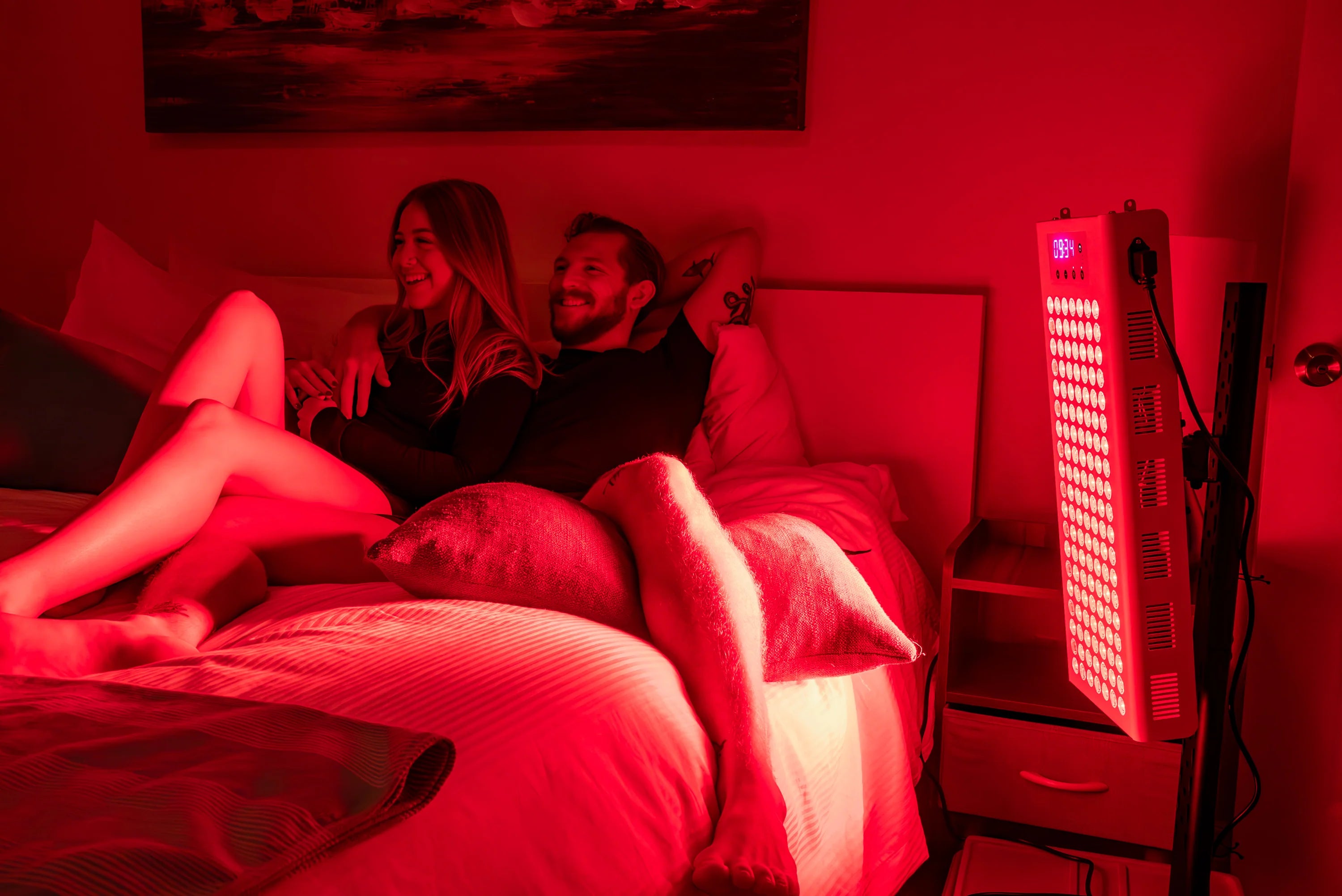 Two people relaxing on a bed beside the RedRush 400 PULSE targeted red and near-infrared light panel