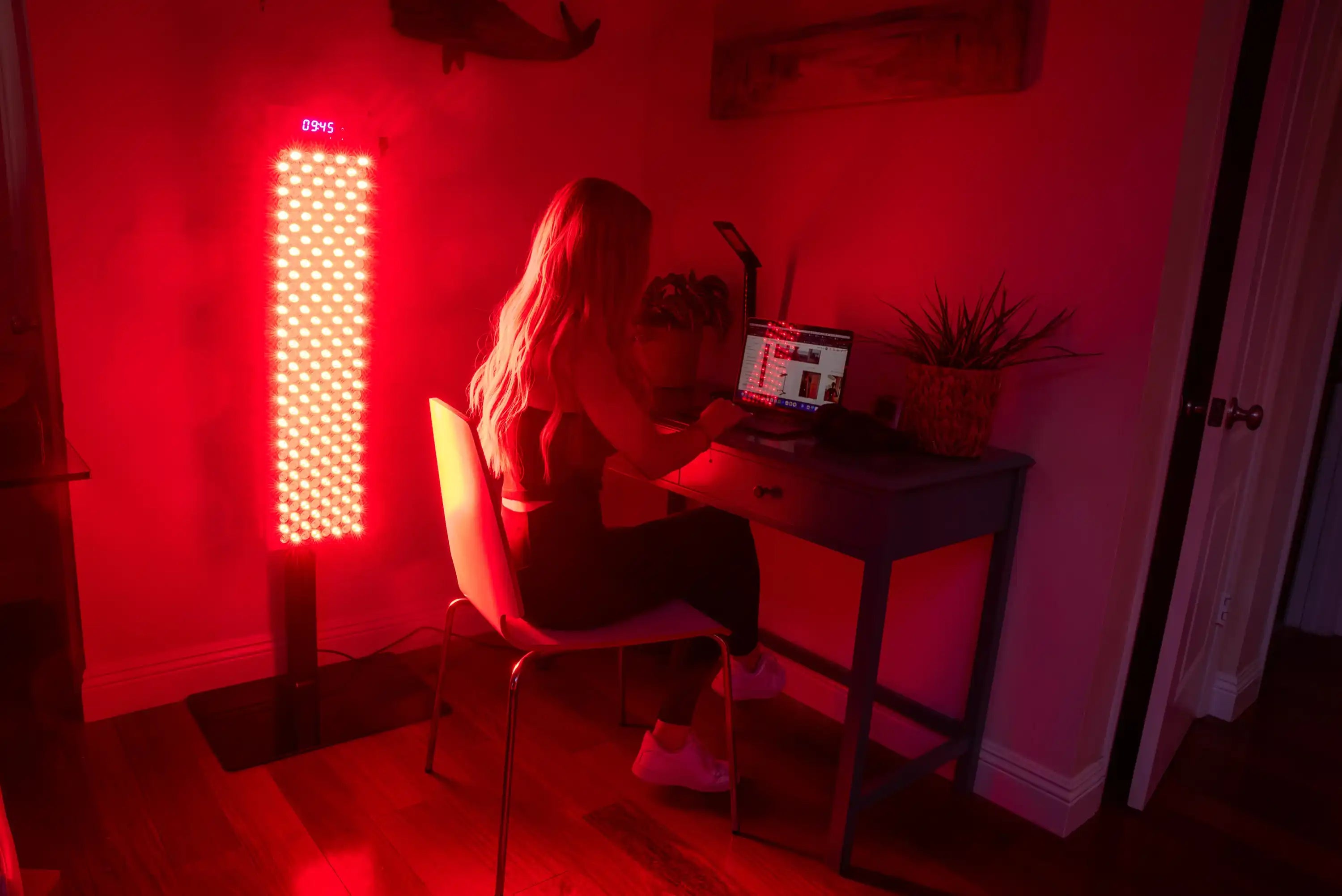 Person working at a desk beside the illuminated RedRush 840 PULSE full-body red and near-infrared light panel